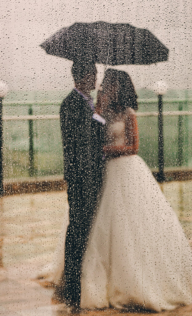 Look from behind the window at wedding couple standing under umbrella in the rain