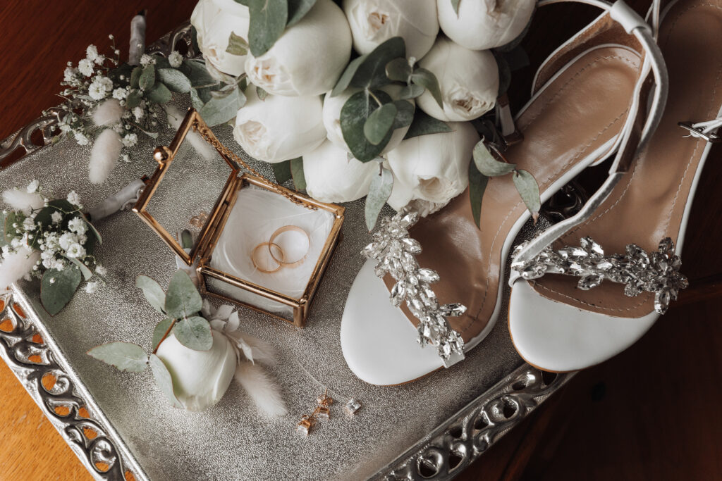 A tray with a white box and a pair of white shoes with a flower on it. The shoes are adorned with jewels and the box contains two rings. Concept of elegance and sophistication